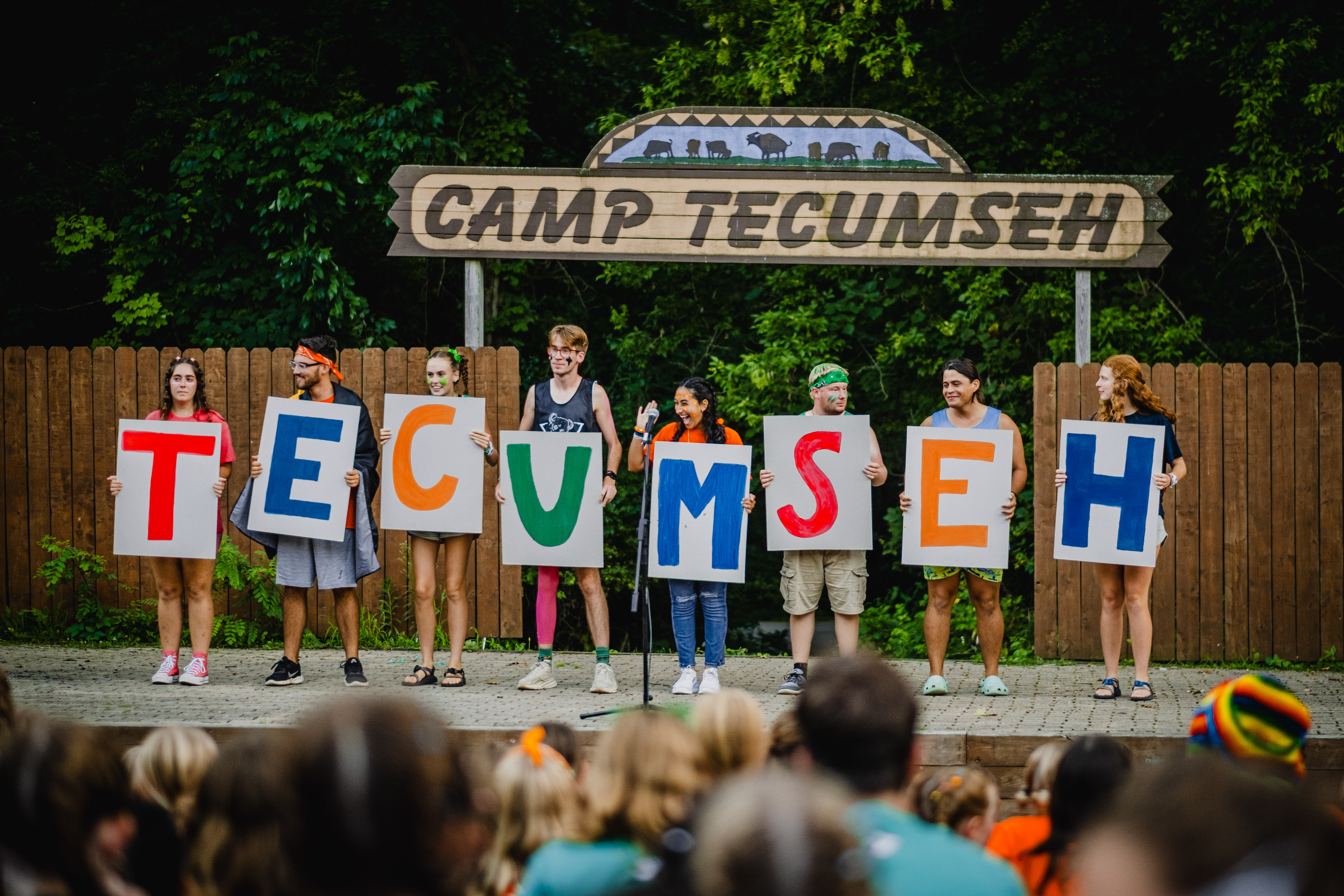Kids from Camp Tecumseh standing in a row and holding signs with colorful painted letters that spell "Tecumseh."