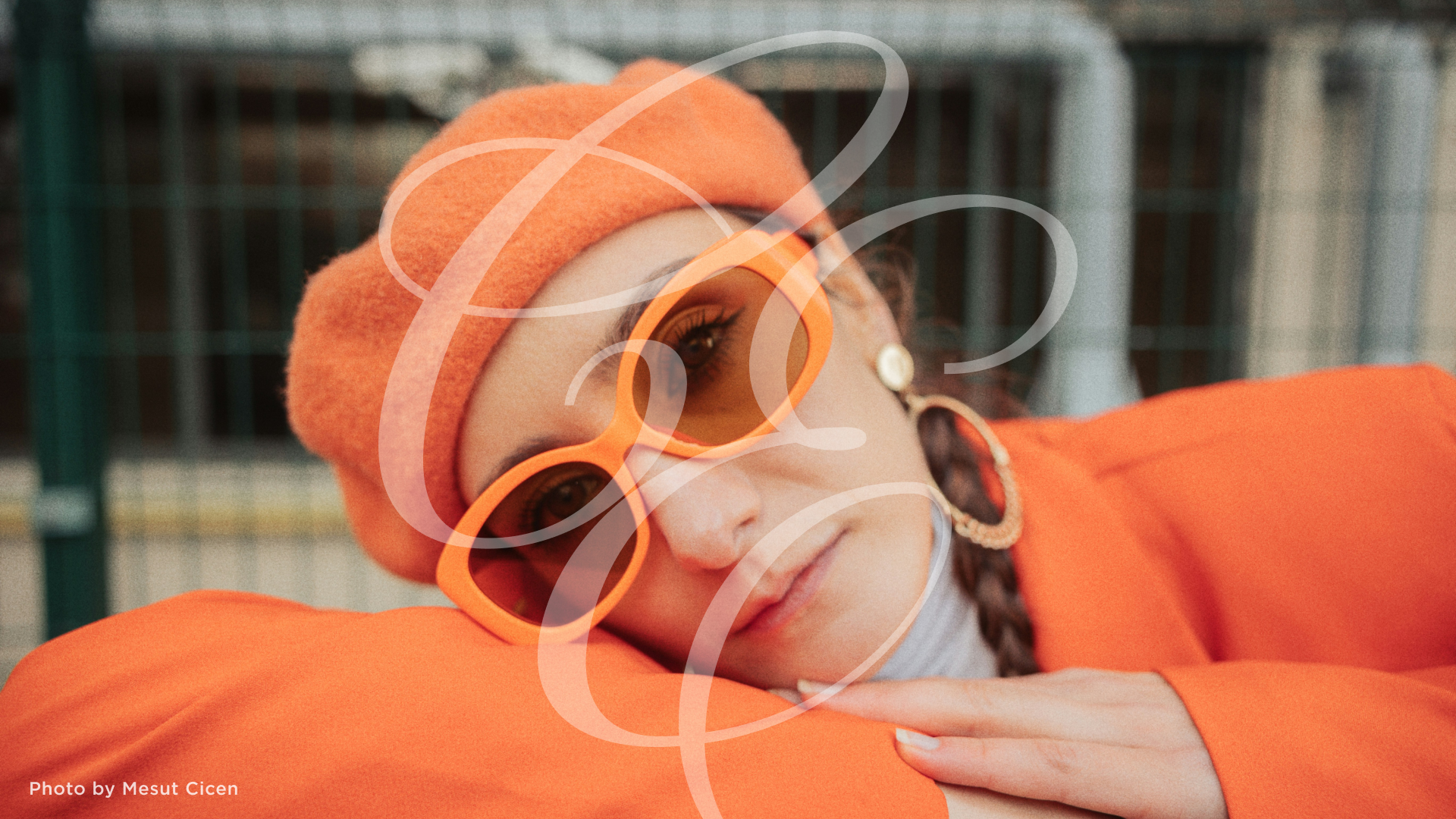 Close-up portrait of a woman wearing an orange sweater, hat, and sunglasses with her cheek lying on crossed arms.