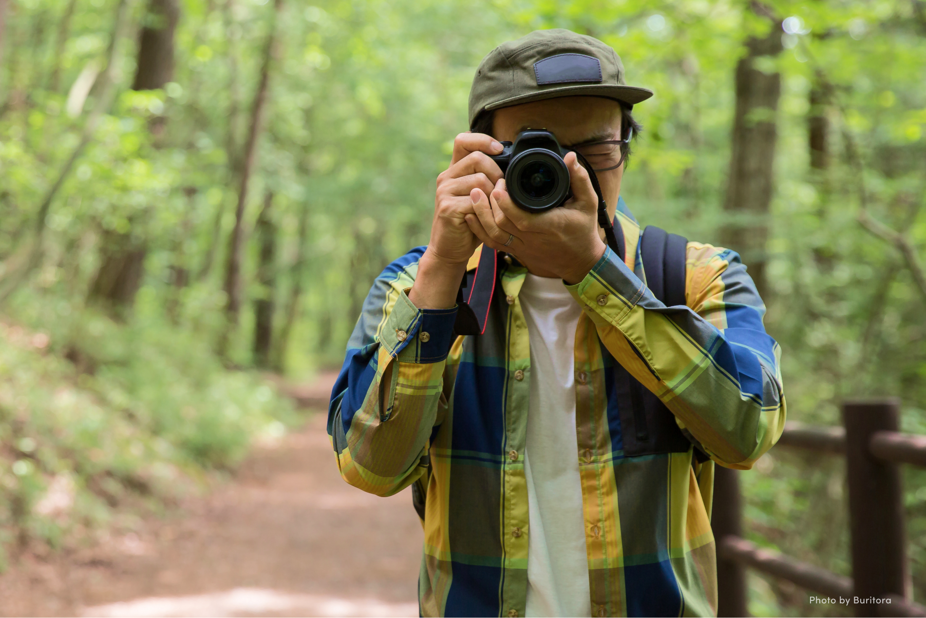 Person with hat, plaid shirt, and glasses pointing a camera at the viewer.